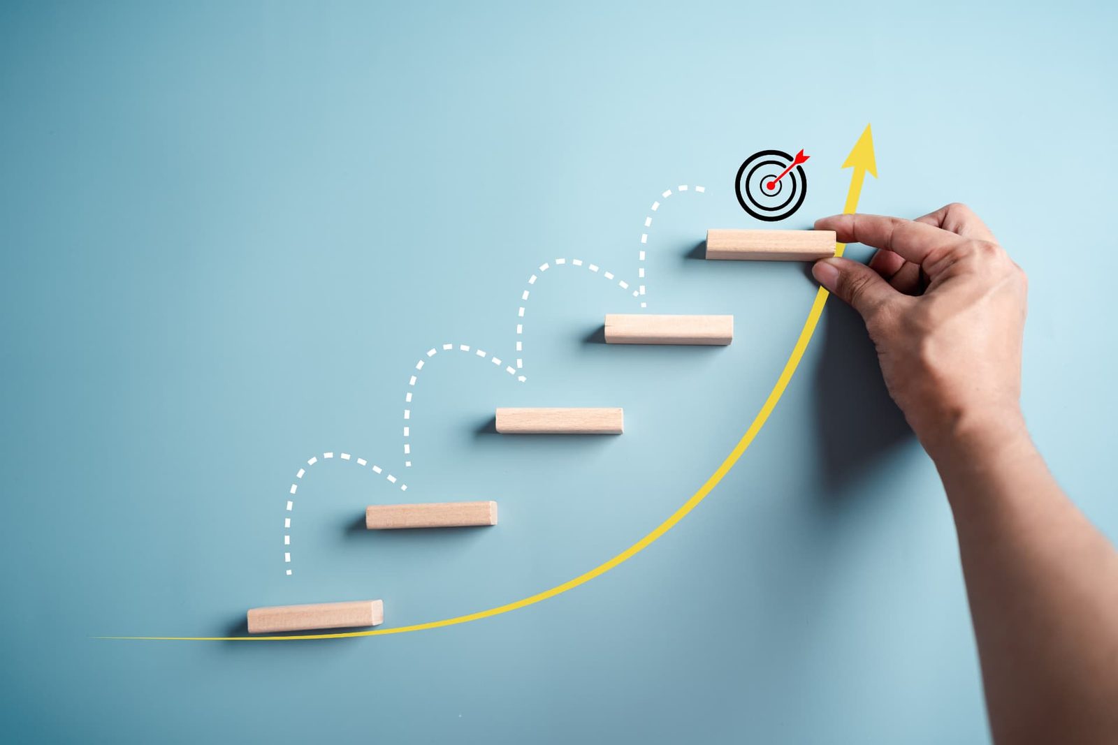A hand placing ascending wooden steps toward a target, alongside a rising arrow
